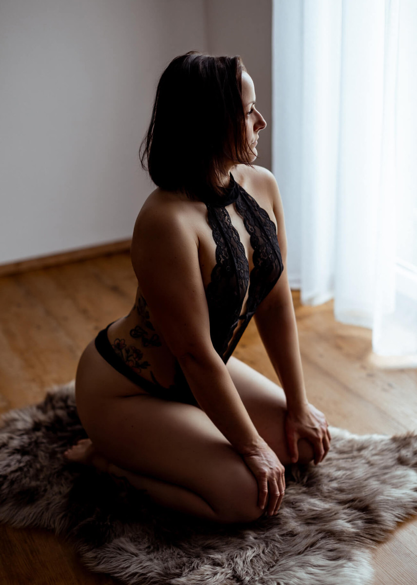 Frau kniet auf Felldecke vor Fenster mit schwarzen Spitzenbody, Erotikfoto